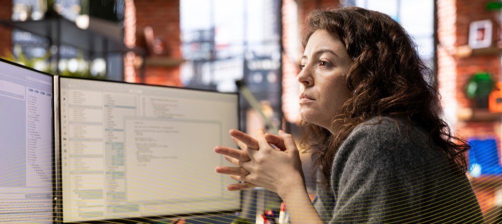 Em ambiente de escritório, uma mulher adulta se debruça sobre uma mesa e analisa dados em uma tela de computador