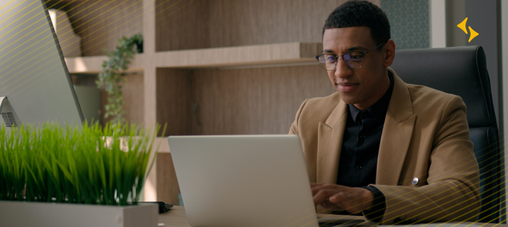 Em ambiente de escritório, homem negro com terno marrom claro utilizando um notebook sobre uma mesa